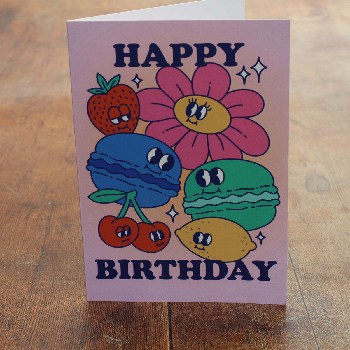 Pink card with colour illustrations of cartoon macarons, fruits and flowers with the text 'happy birthday'