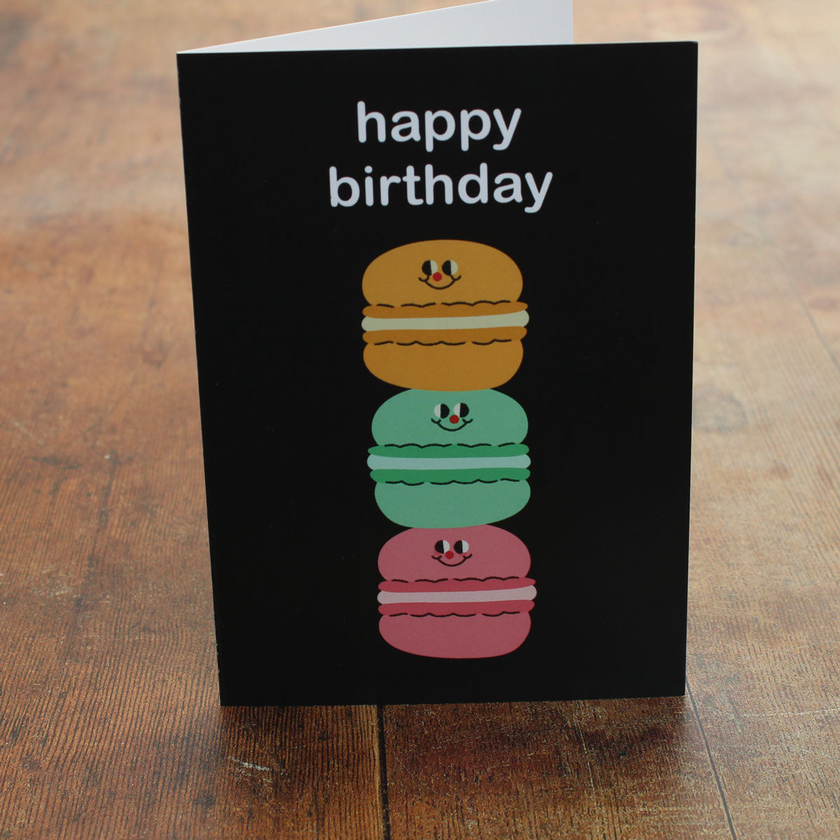 Black birthday card with colourful macaron illustration of three macarons stacked on top of one another and 'happy birthday' text on a wooden surface