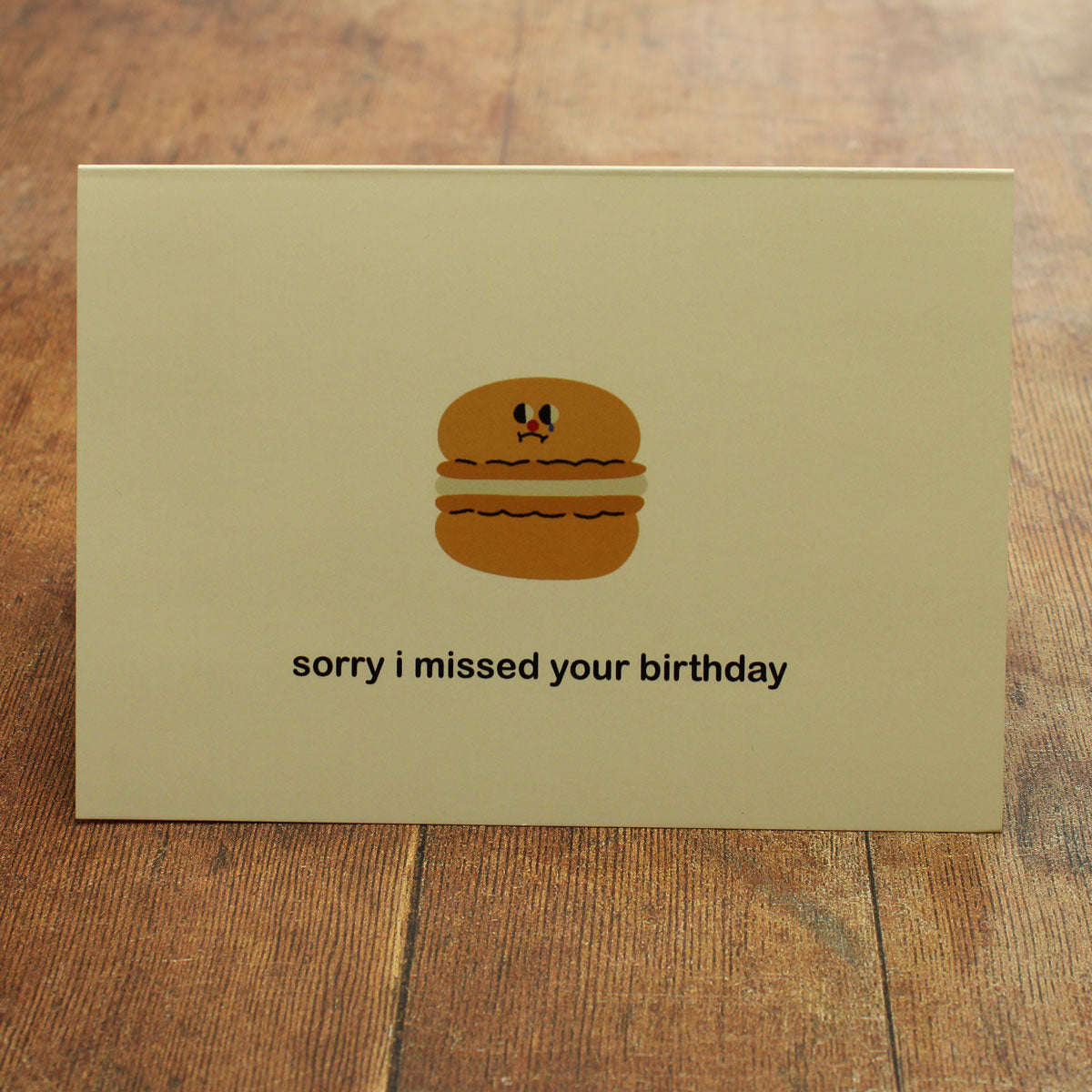 Cream greeting card with a cartoon macaron and 'sorry I missed your birthday' text on a wooden surface