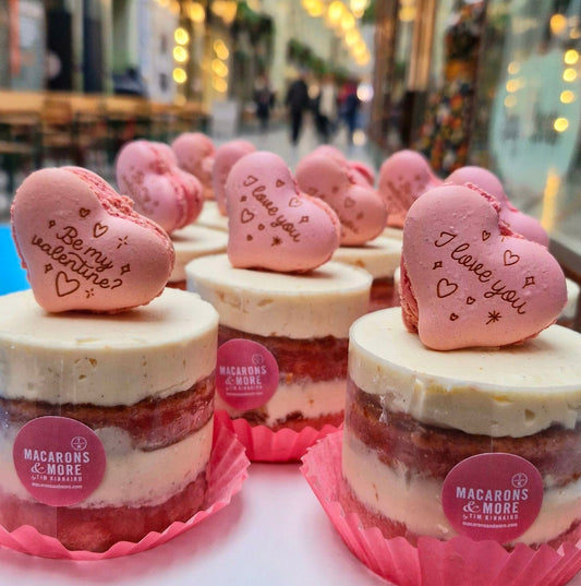 Mini layered cakes with pink engraved macaron hears on top, in a shop window