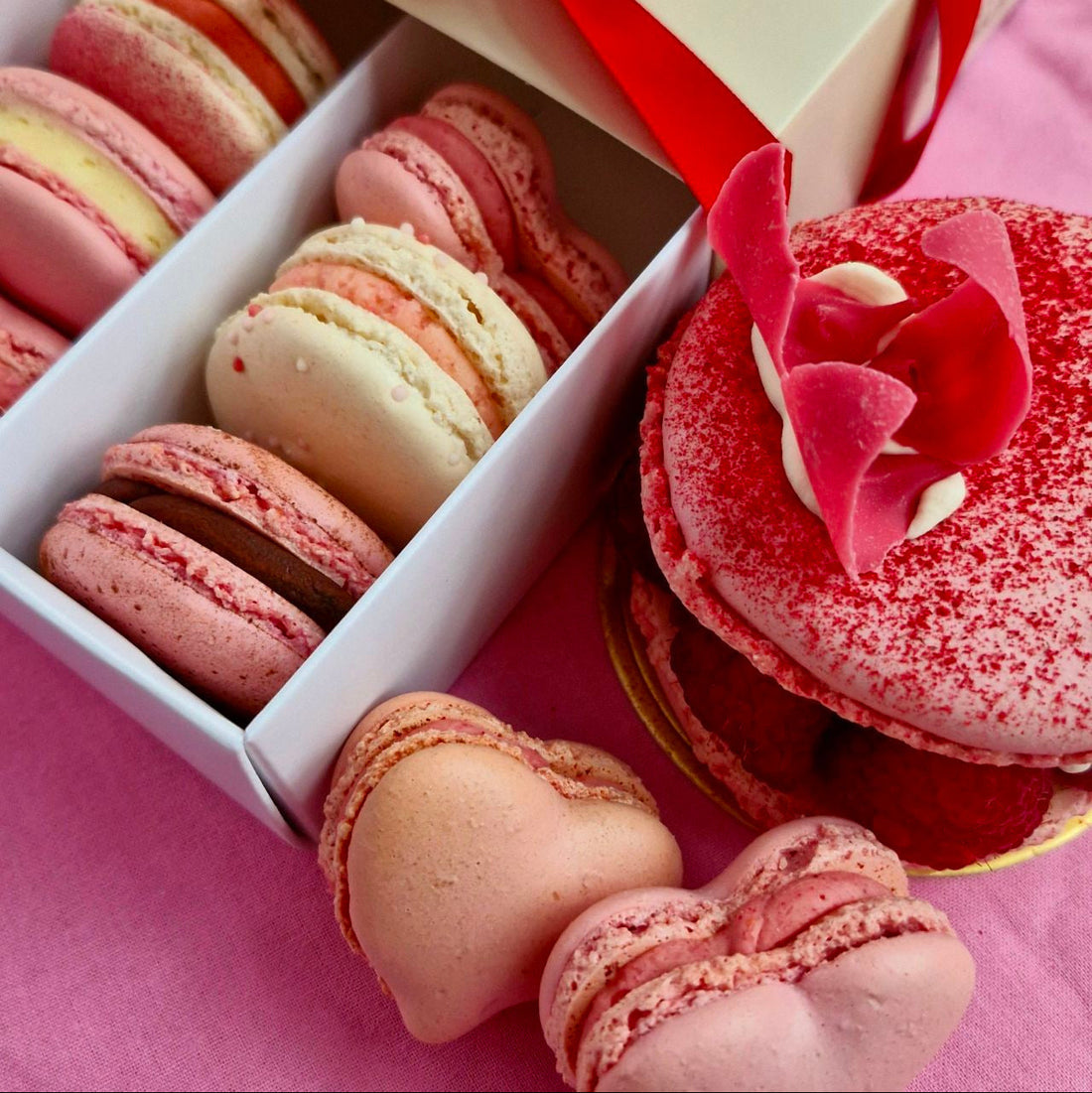 Variations of pink and cream macarons, some inside a white box, next to a larger macaron on a pink surface
