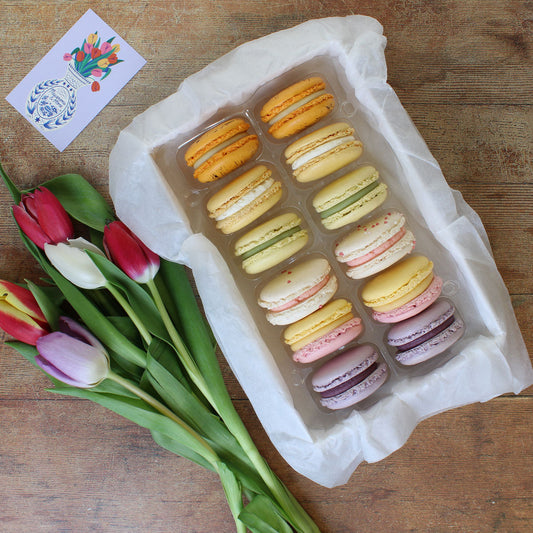 Box of colour macarons next to a bunch of tulips and a small postcard with a flower illustration, on a wooden surface.