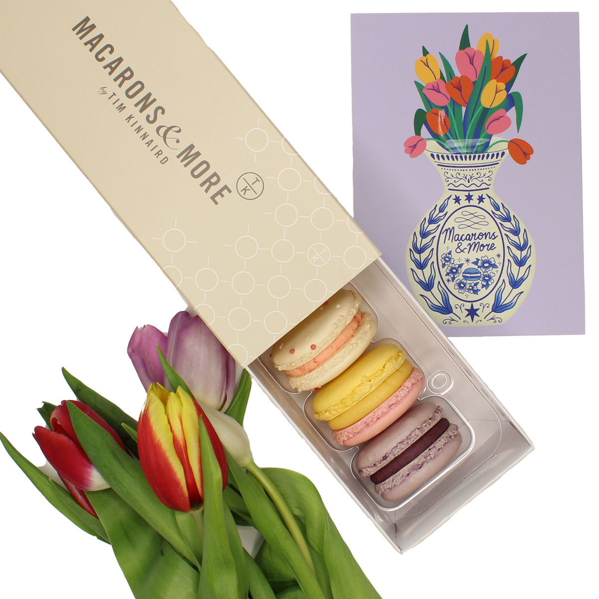 Box of macarons next to tulips and a decorative card on a white background