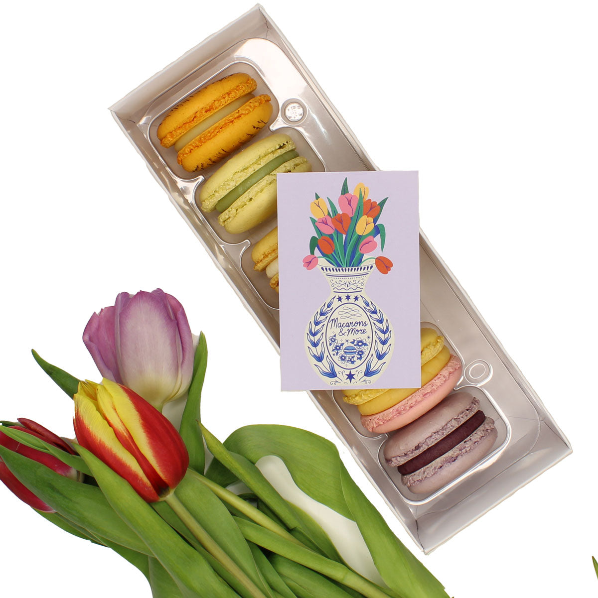 Box of colorful macarons with a card featuring a floral design, accompanied by tulips on a white background.