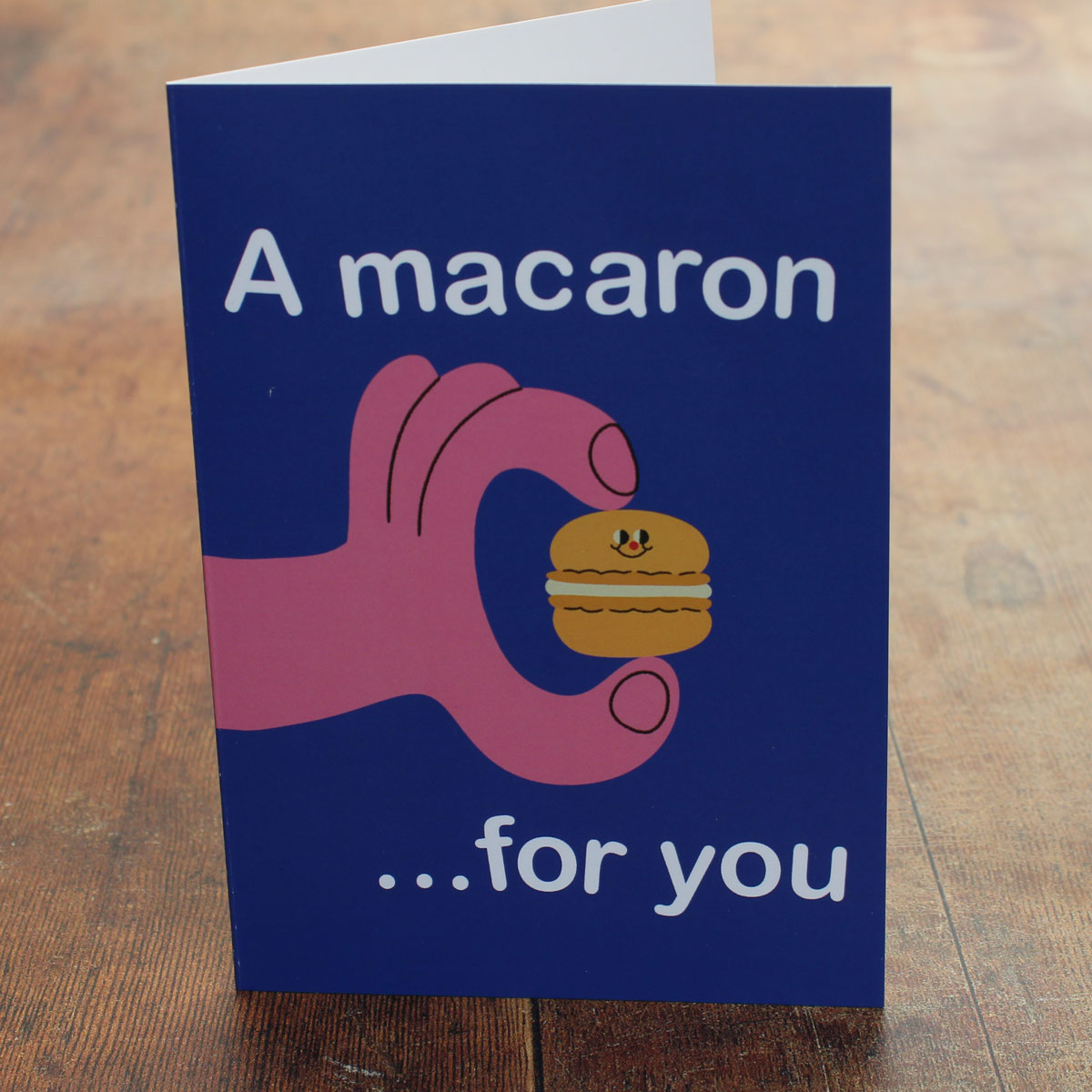 Card with a hand holding a macaron and text 'A macaron ...for you' on a wooden surface