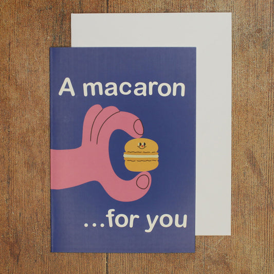 Card with a hand holding a macaron and text 'A macaron ...for you' on a wooden surface.