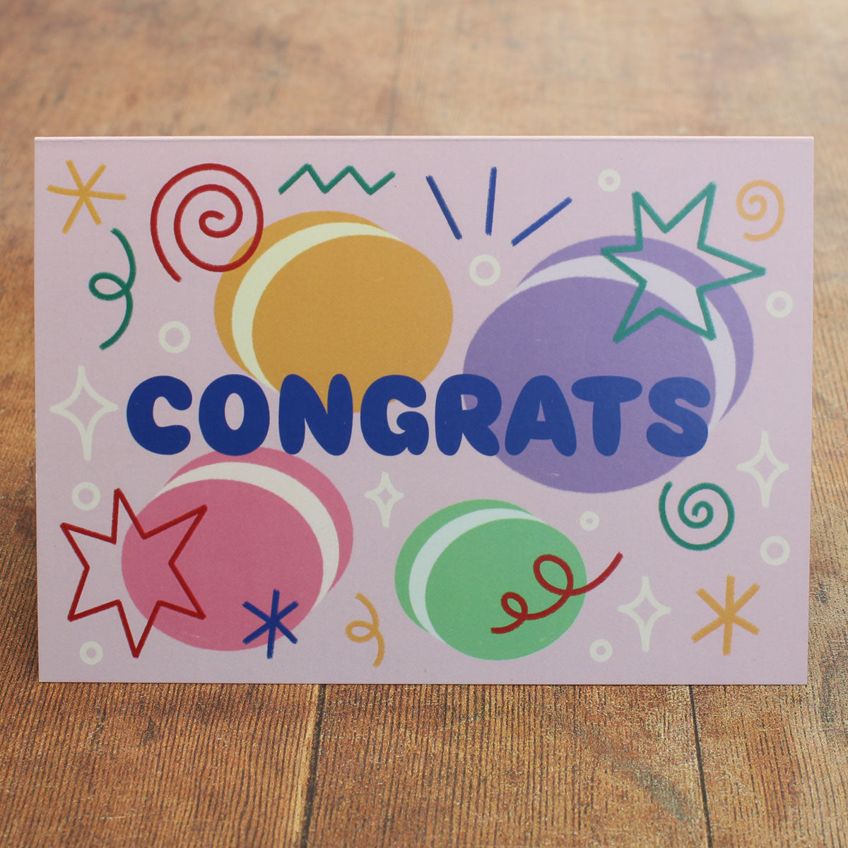 Colourful card with 'Congrats' text with macaron illustrations and stars and swirls on a wooden surface