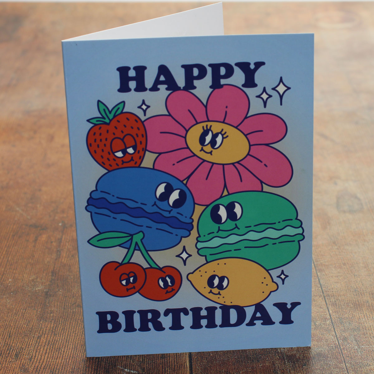 Blue card with colour illustrations of cartoon macarons, fruits and flowers with the text 'happy birthday'