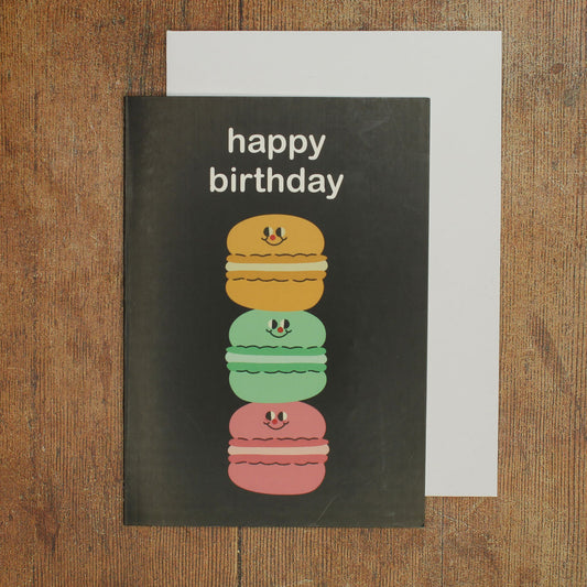 Black birthday card with colourful macaron illustration of three macarons stacked on top of one another and 'happy birthday' text on a wooden surface