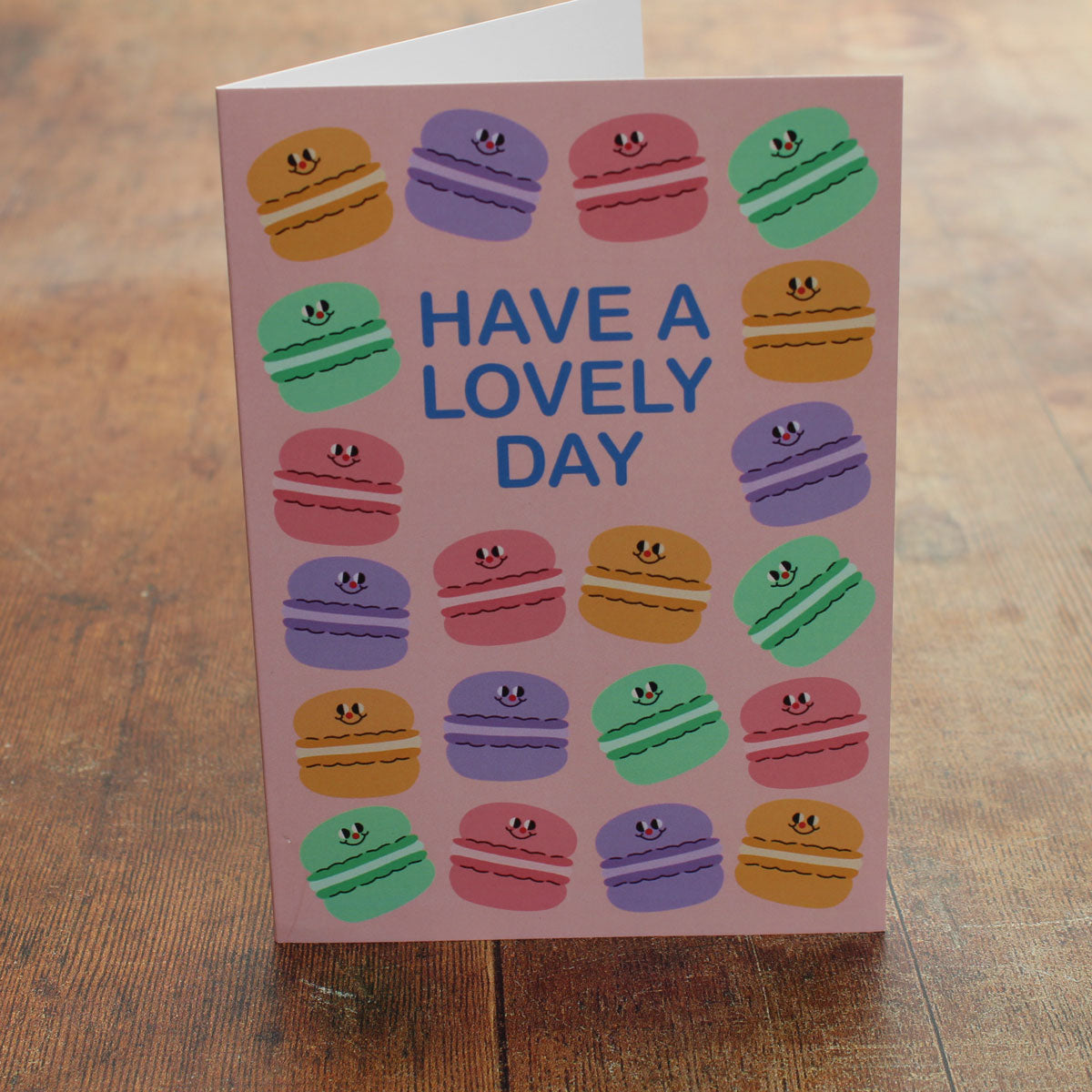 Greeting card with colourful macaron illustrations on a pink background and 'Have a Lovely Day' text on a wooden surface
