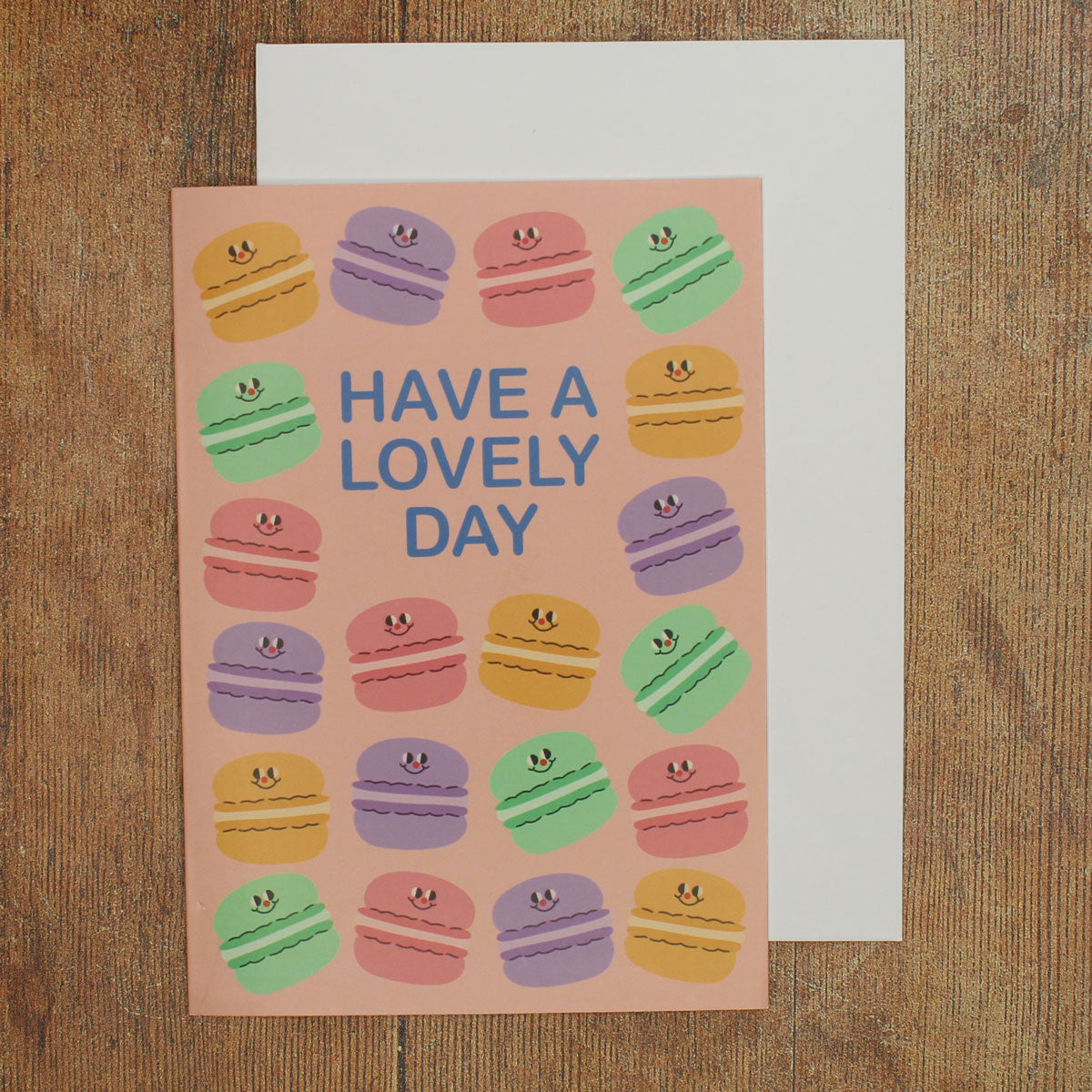 Greeting card with colourful macaron illustrations on a pink background and 'Have a Lovely Day' text on a wooden surface