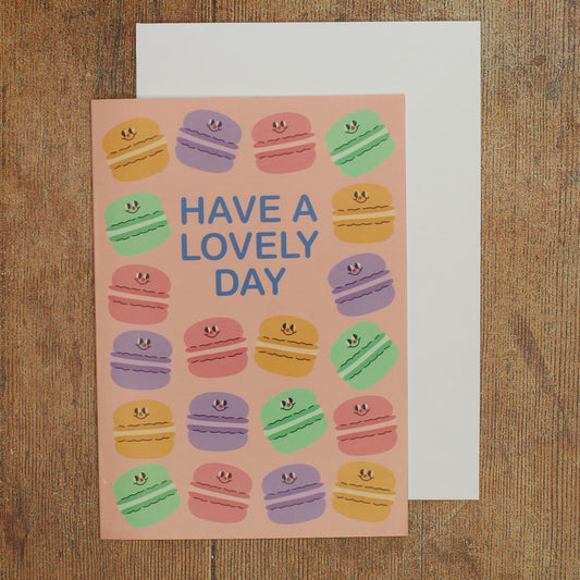 Greeting card with colourful macaron illustrations on a pink background and 'Have a Lovely Day' text on a wooden surface
