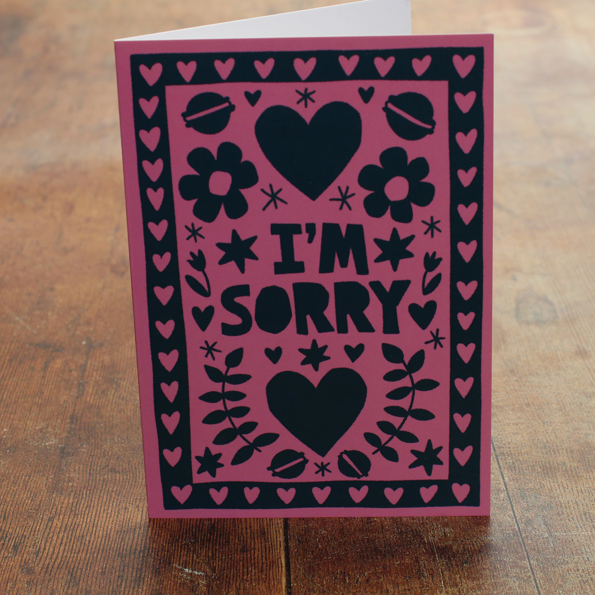 Card with 'I'm Sorry' text on a pink background with black hearts and flowers and macarons on a wooden surface