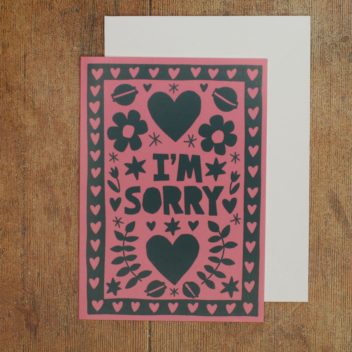 Card with 'I'm Sorry' text on a pink background with black hearts and flowers and macarons on a wooden surface
