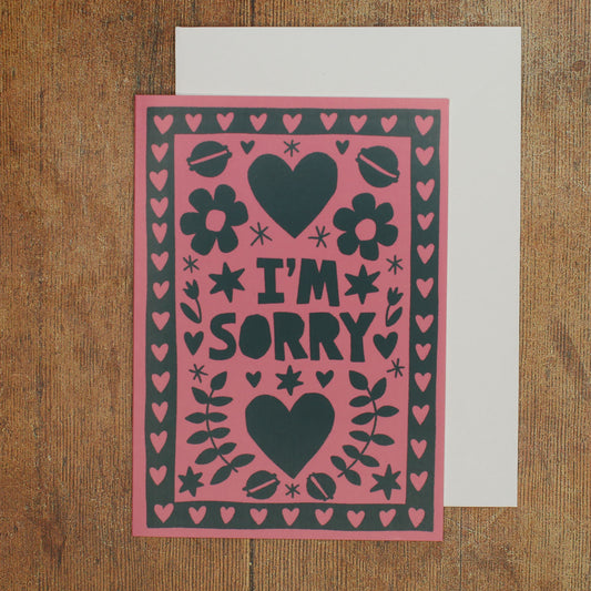 Card with 'I'm Sorry' text on a pink background with black hearts and flowers and macarons on a wooden surface