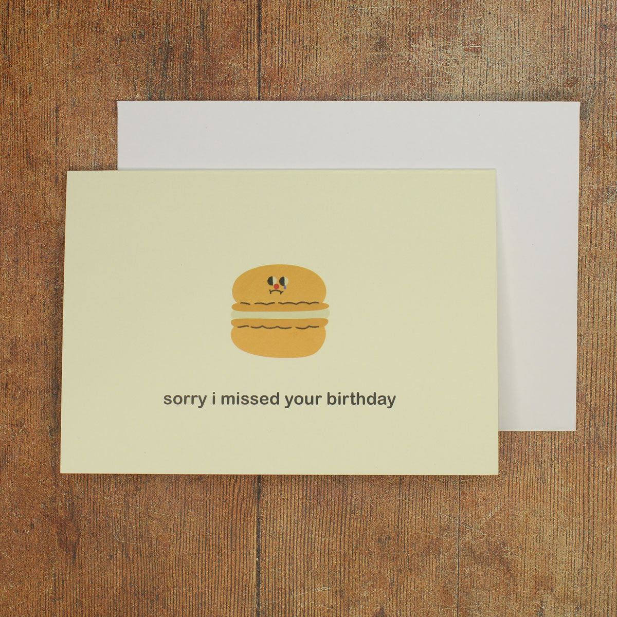 Cream greeting card with a cartoon macaron and 'sorry I missed your birthday' text on a wooden surface