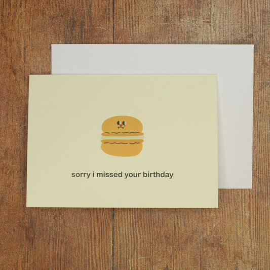 Cream greeting card with a cartoon macaron and 'sorry I missed your birthday' text on a wooden surface