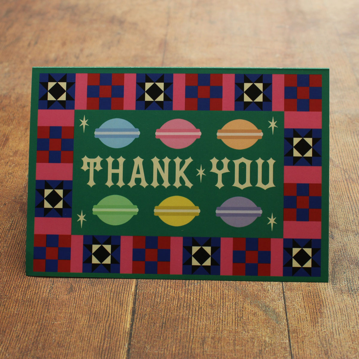Green card with colourful  macaron illustrations and a colourful border with 'Thank You' text  on a wooden surface