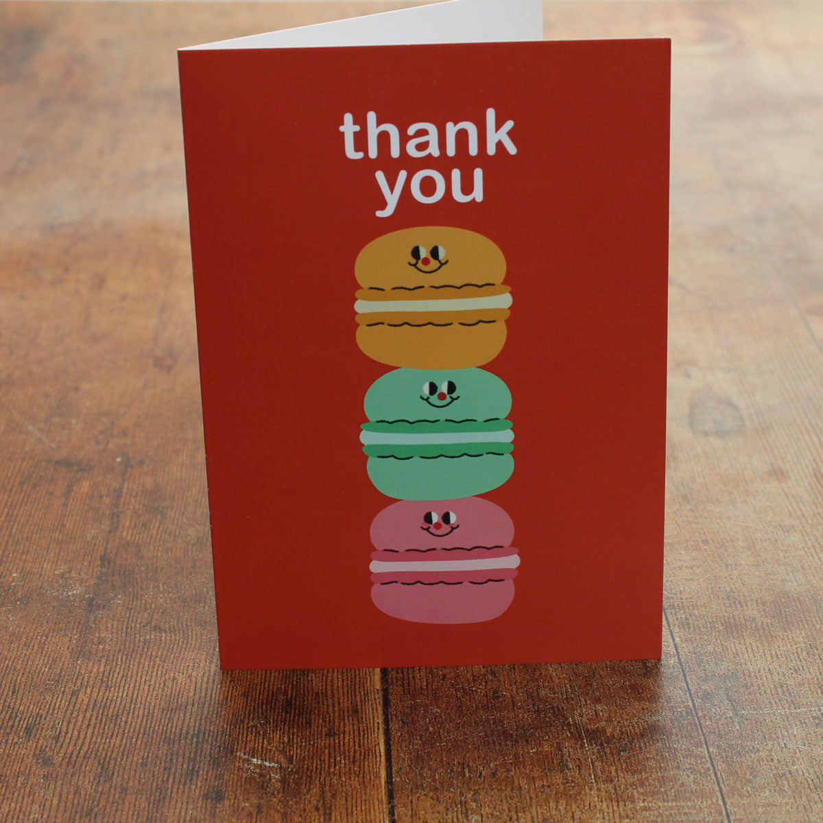 Orange greeting card with colorful illustration of a stack of macarons and 'thank you' text on a wooden surface