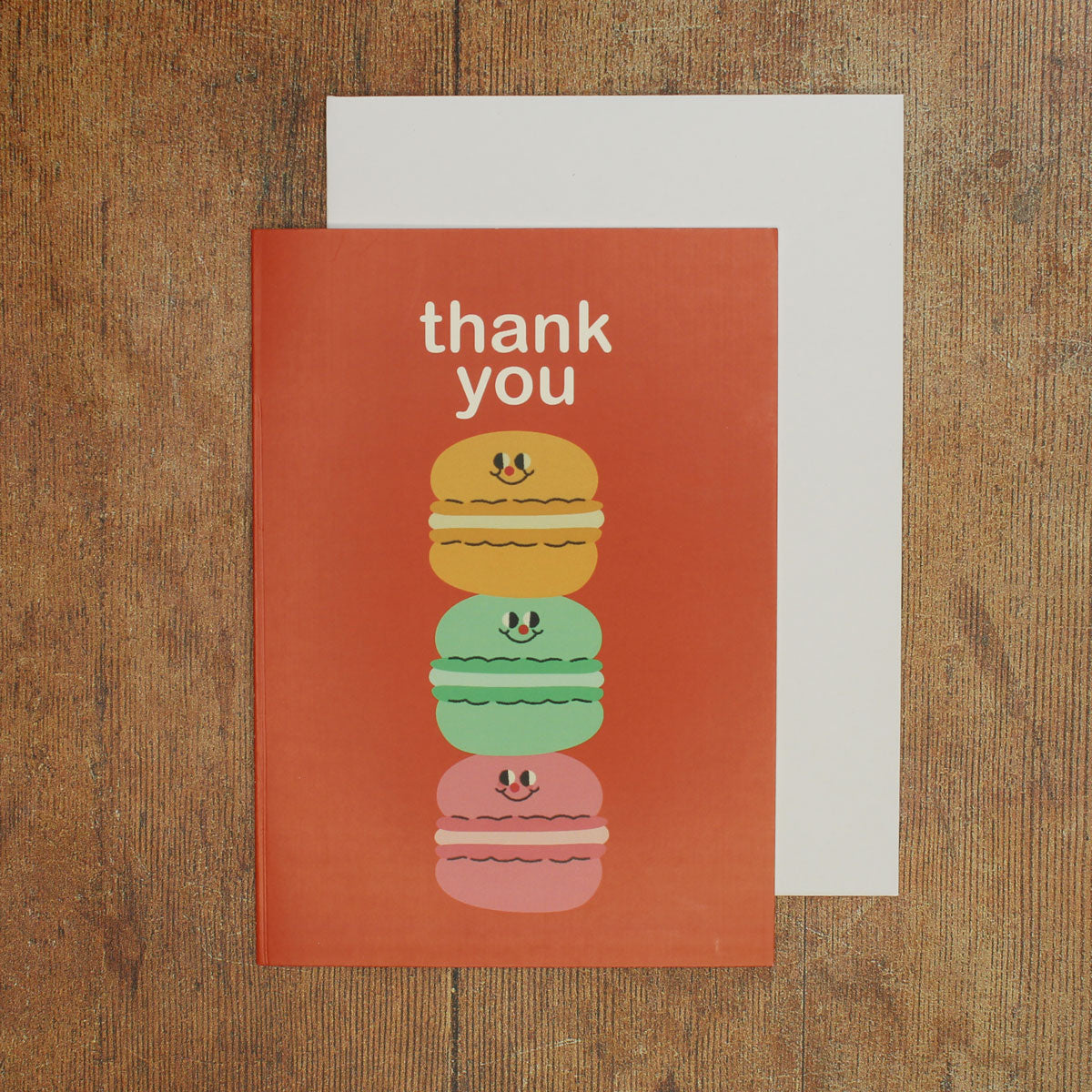 Orange greeting card with colorful illustration of a stack of macarons and 'thank you' text on a wooden surface
