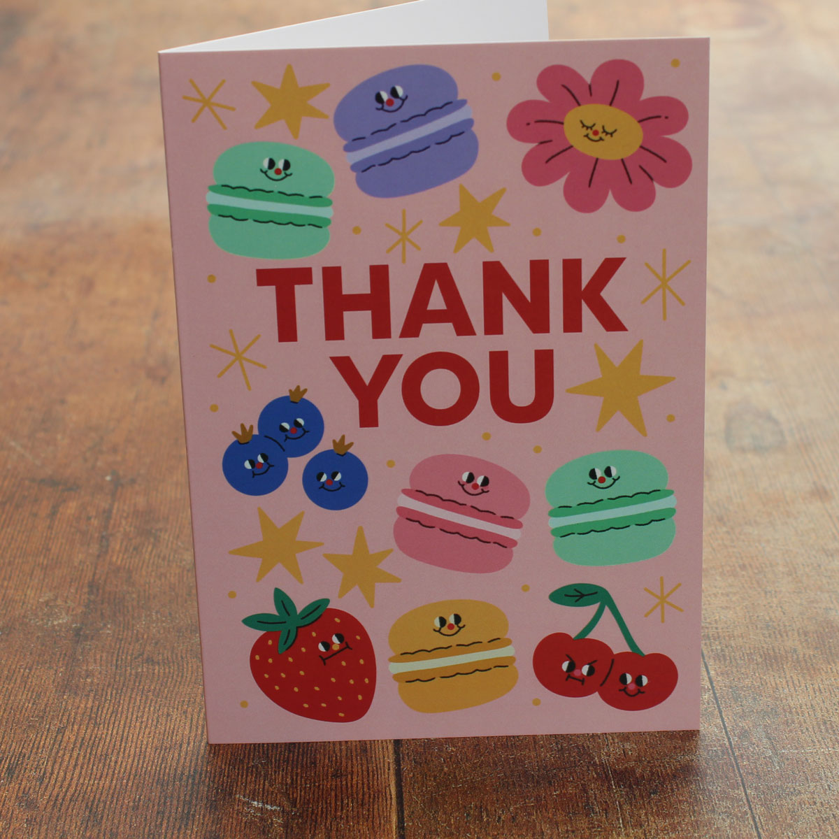 Pink card with colorful illustrations of macarons, fruits, flowers and stars, with 'Thank You' text on a wooden surface