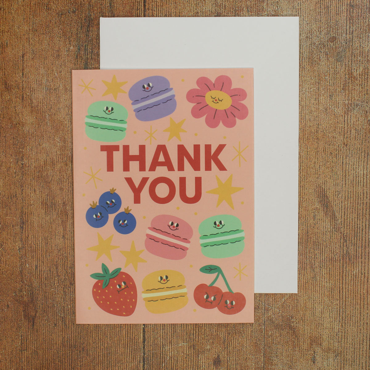 Pink card with colorful illustrations of macarons, fruits, flowers and stars, with 'Thank You' text on a wooden surface