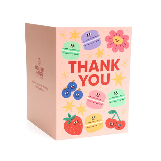 Pink card with colorful illustrations of macarons, fruits, flowers and stars, with 'Thank You' text on a wooden surface