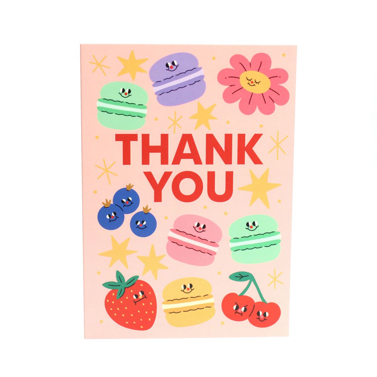 Pink card with colorful illustrations of macarons, fruits, flowers and stars, with 'Thank You' text on a wooden surface