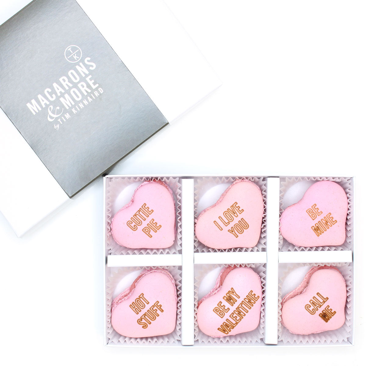 Heart-shaped pink macarons with engraved valentines messages, in a box labeled 'Macarons & More'.